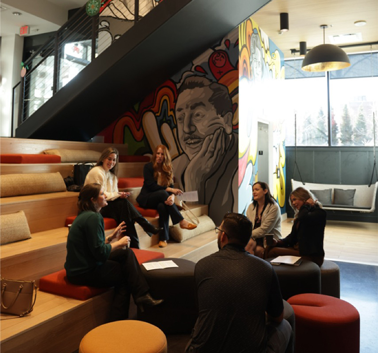 A group of people sits on a set of lecture stairs and ottomans.  