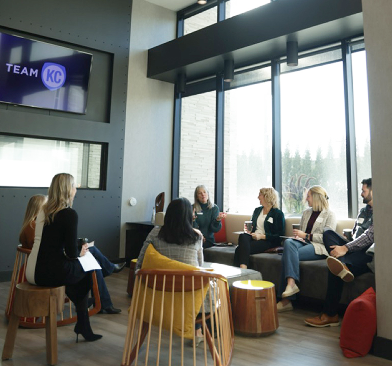 A group of individuals gathers around a set of small tables, discussing papers in their hands. 