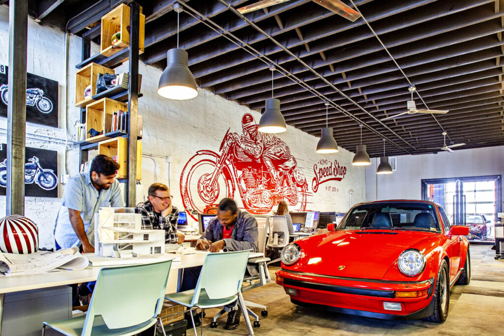 Three men chat at a table next to a red sports car indoors.