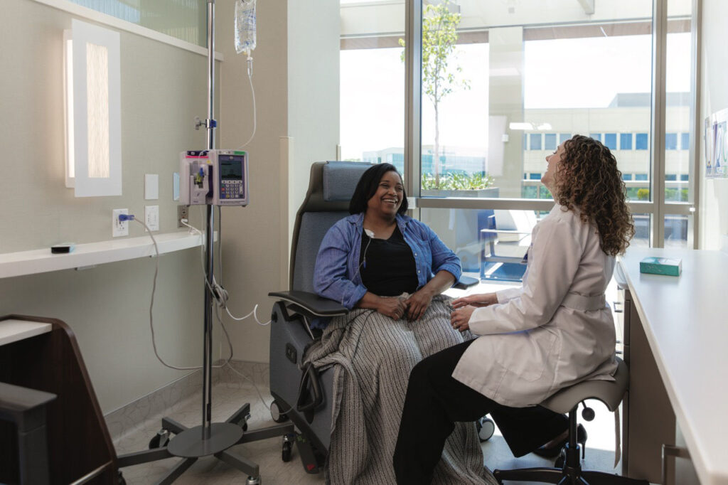 Female doctor speaking to a female patient seated and hooked up to an IV, both are laughing