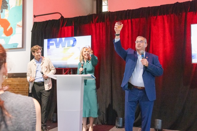 A man cheers the crowd from a stage, joined by a man to the left and a woman in the middle.