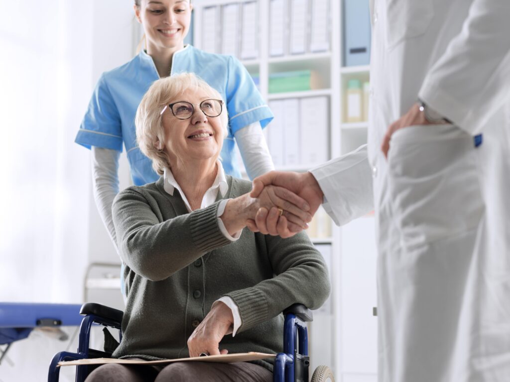 Elderly woman shaking hands with a doctor out of frame, being pushed in a wheelchair by a young female nurse