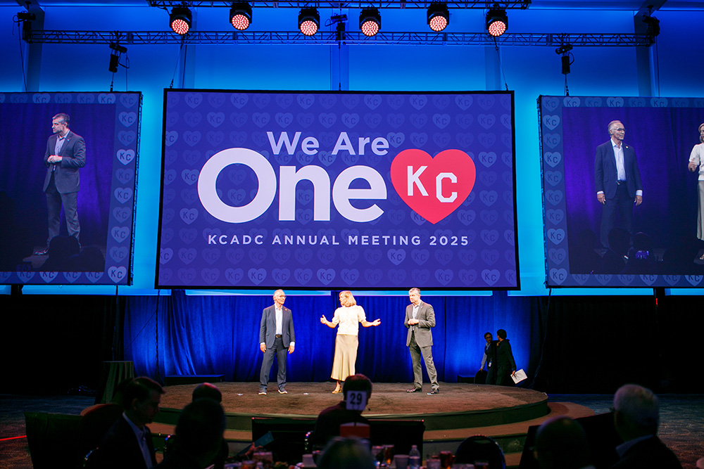 Two men and a woman standing on a stage in front of a large screen that reads We Are OneKC