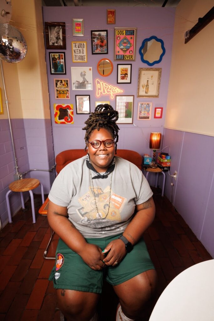 Black woman in casual dress sitting in front of a purple wall with eclectic art