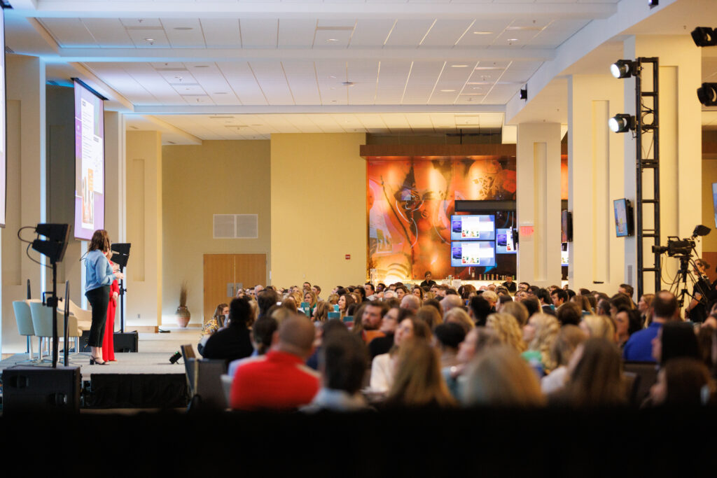 Two women standing on a stage in front of hundreds of people seated at tables