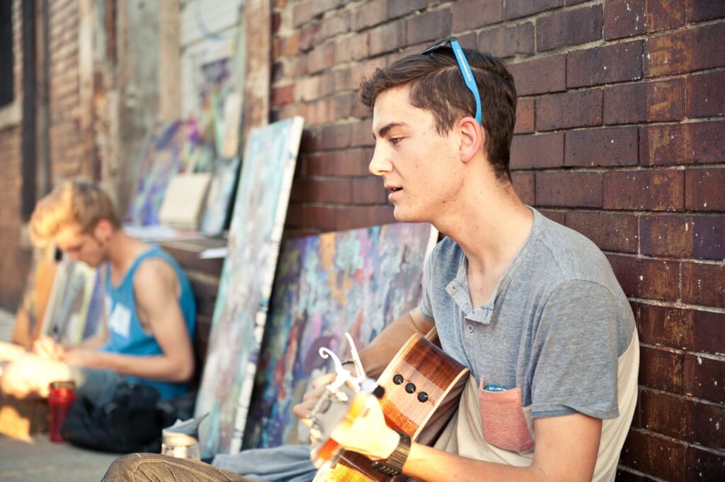 Young man seated against a brick wall playing a guitar