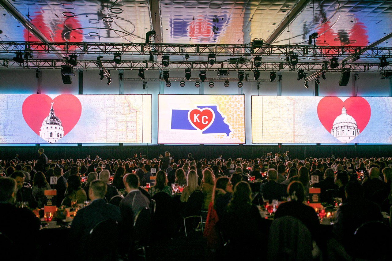 Large room full of people seated at tables with large screens displaying OneKC