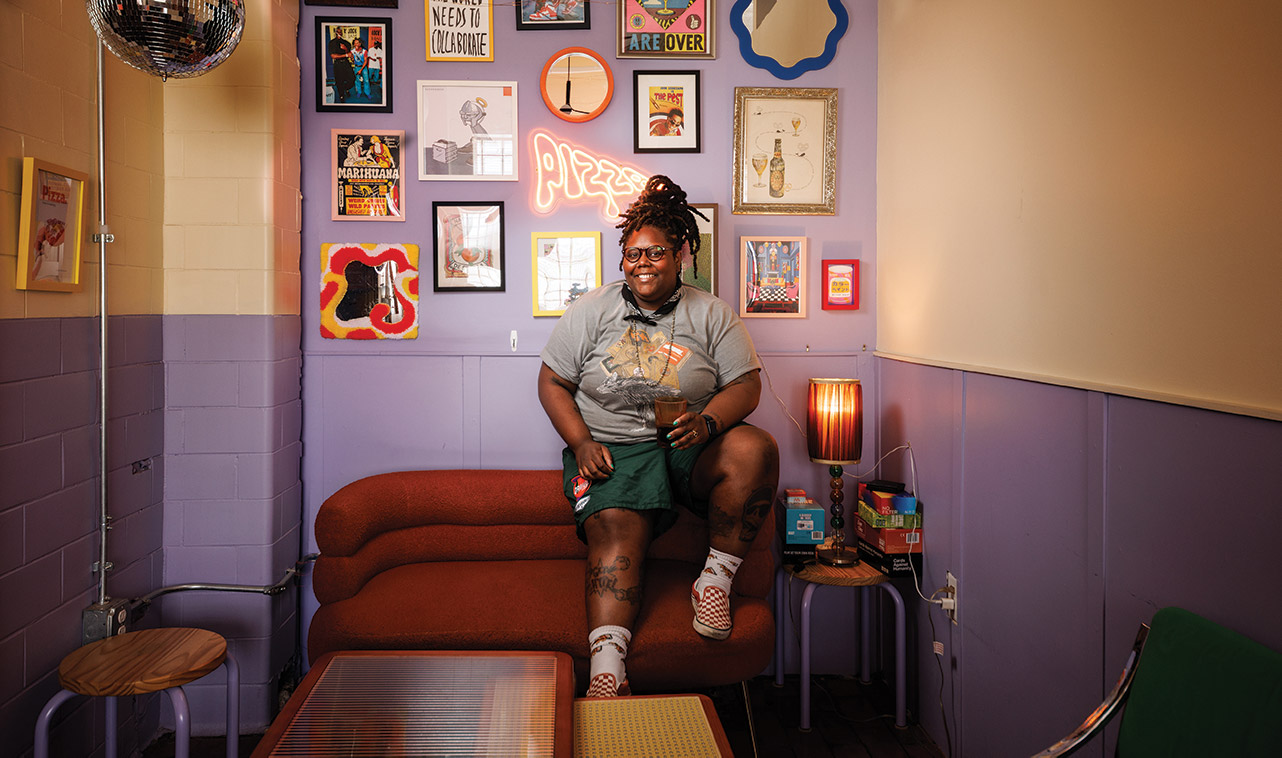 Jhy Coulter sitting on a couch inside with a gallery wall behind her.