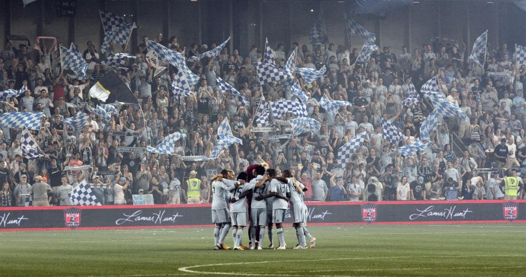 Men's soccer team player standing huddled on a field with a lively crowd cheering behind them