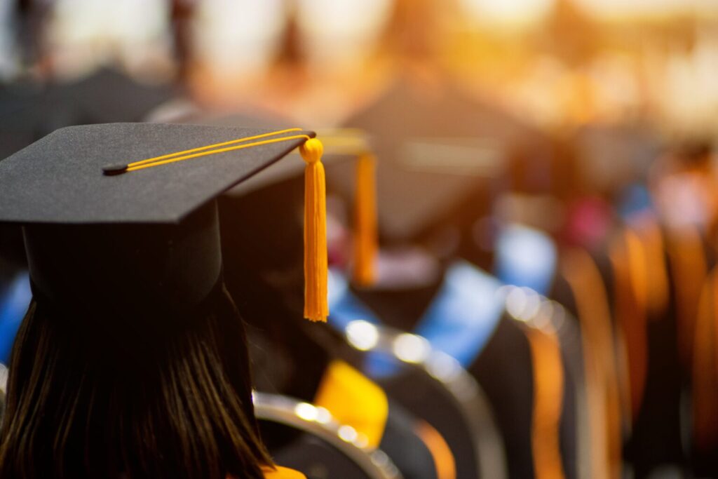 Back of dark haired person wearing a black graduation cap with a yellow tassel, with more graduates blurry in the background