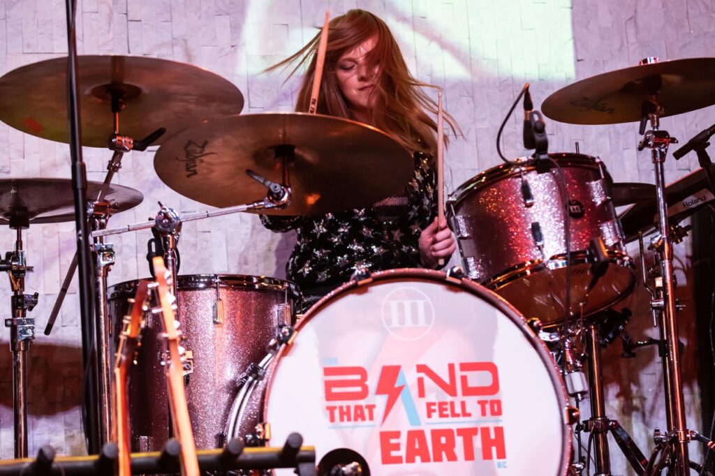 Girl playing behind a drumset, with Band That Feel to Earth written on the bass drum