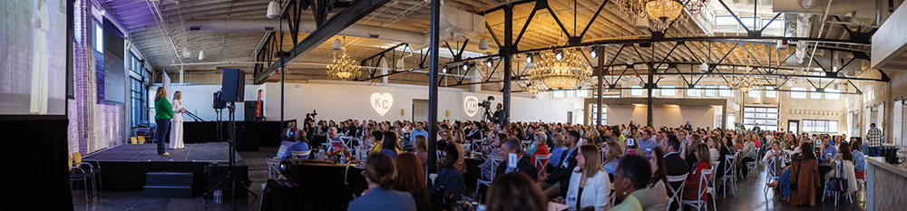Wide shot of two women on stage addressing hundreds of seated attendees in a large, chic industrial event space