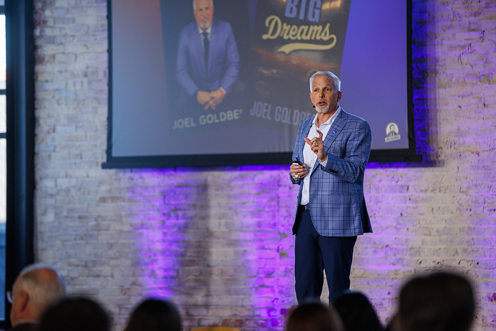 Joel Goldberg in a blue plaid suit standing on a stage addressing a crowd
