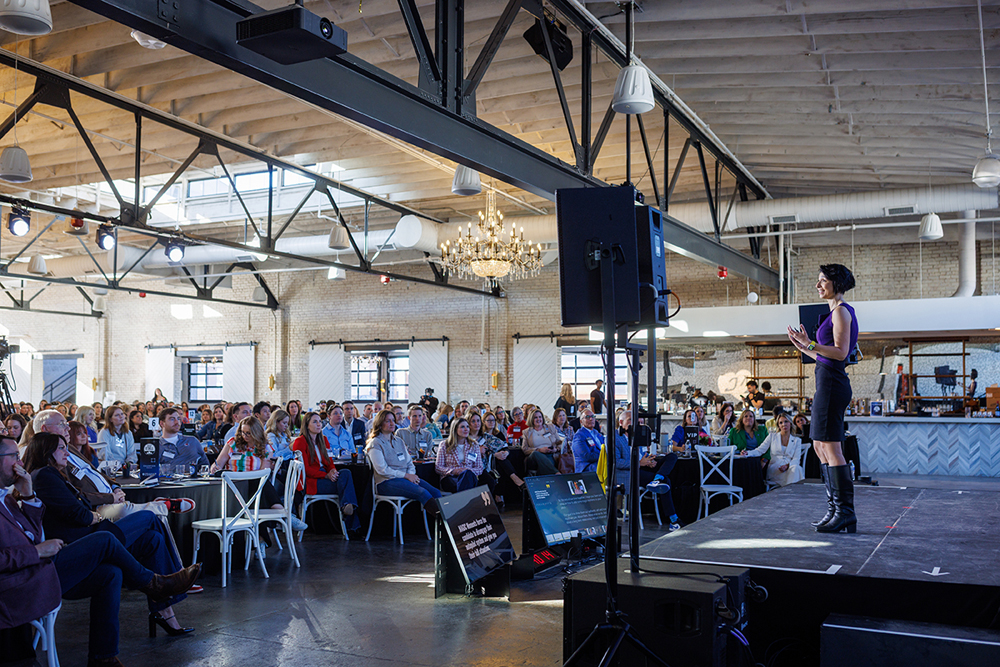 Diana Kander standing on stage addressing hundreds of seated attendees in a large, chic industrial event space
