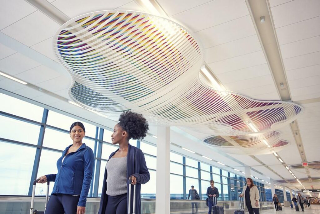 Travelers with suitcases walking through a large hallway with a rainbow cloud-like art installation on the ceiling
