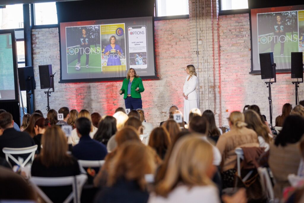 Two women present from a stage, viewed from the audience.