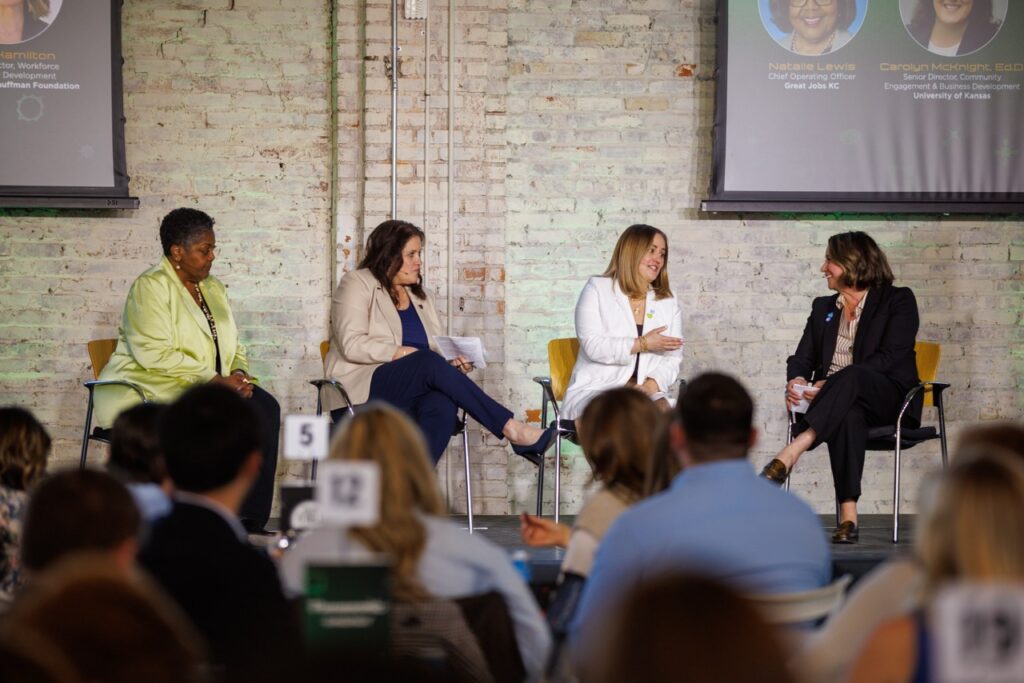 Four women sit in chairs on a stage, speaking out to an audience. 