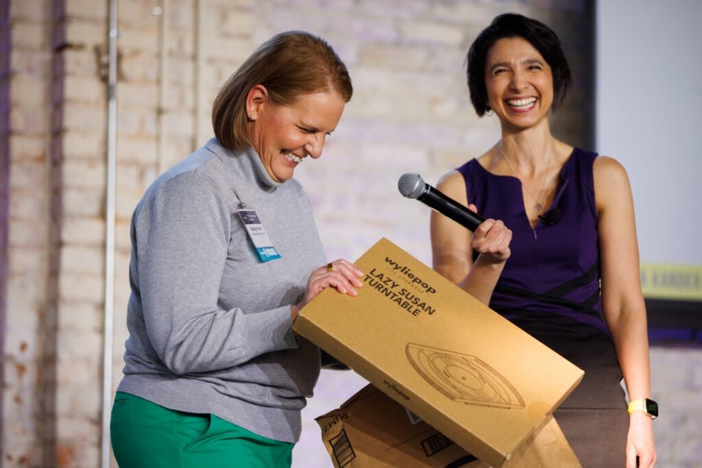 Woman opens an amazon package from stage to fins a refrigerator lazy suzan. Woman sstands next to her with a microphone laughing.