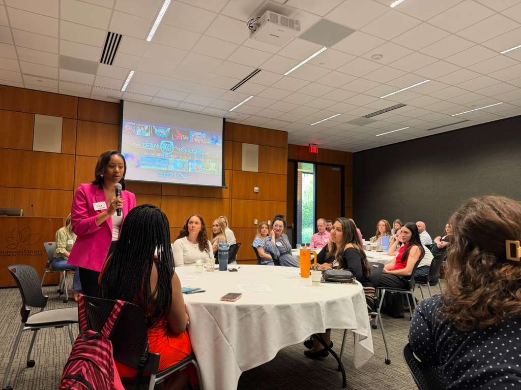 Woman in a pink jacket speaks to a room of people sitting at roundtables.