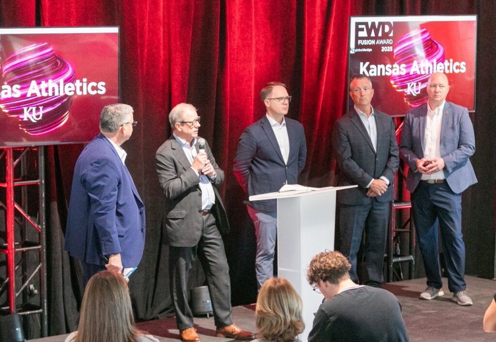 Five men in suits stand on a stage with Kansas Athletics displayed on screens behind them