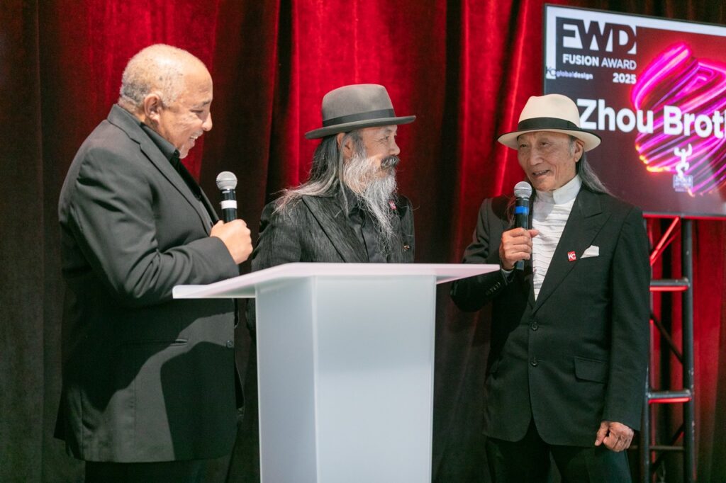 Three men in suits stand on a stage holding microphones, with Zhou B Arts Center displayed on a screen behind them