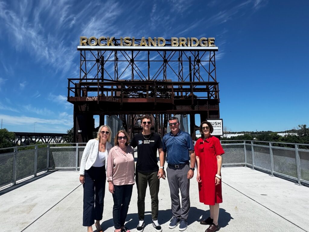 Several people standing in front of the Rock Island Bridge sign, with a blue sky in the background.