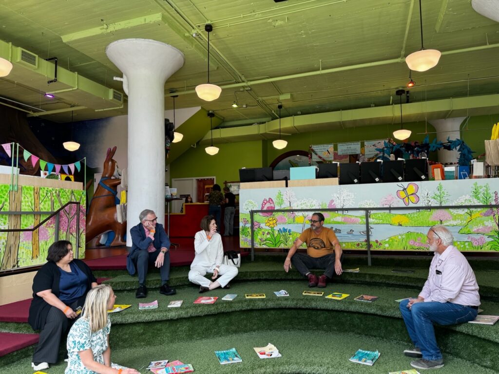 Several people sitting on a carpeted tiered seating area, surrounded by children's books.