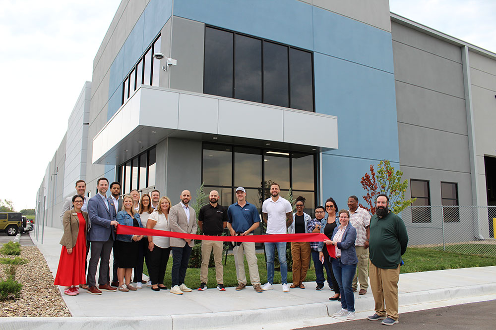 Group of people cutting a large red ribbon in front of an industrial building