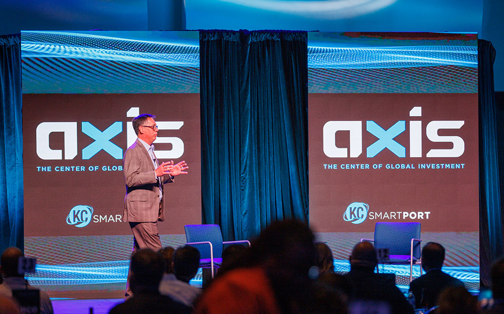 Man standing on stage with LED banners showing AXIS The Center of Global Investment, KC SmartPort